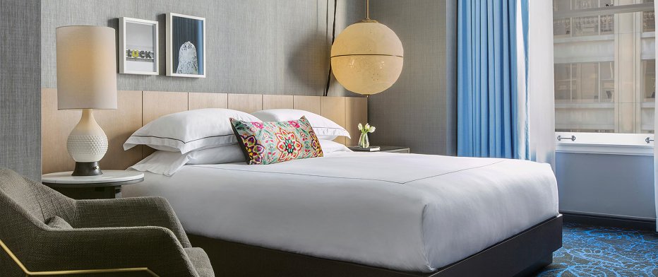 Hotel bedroom with white bedding, floral accent pillow, and a round paper pendant light beside a window with blue curtains