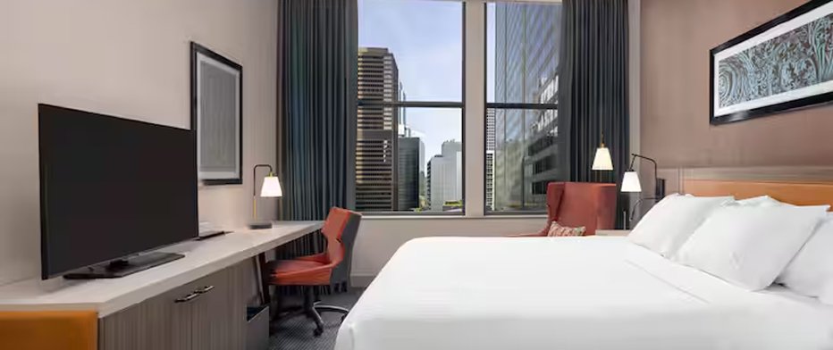 Hotel bedroom with a wall-mounted TV, work desk with orange chair, and a floor-to-ceiling window overlooking a city skyline