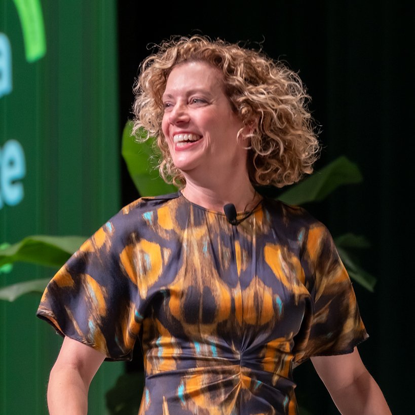 A woman with curly hair laughing on stage while speaking at a conference