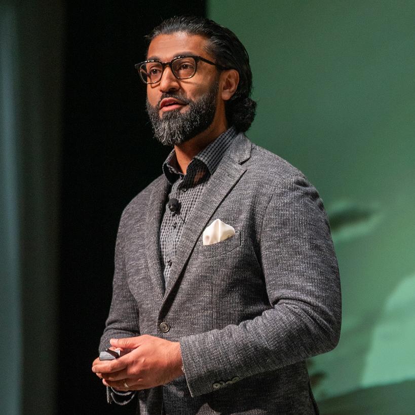 A bearded man in glasses and a gray blazer speaking on stage with a green-lit background