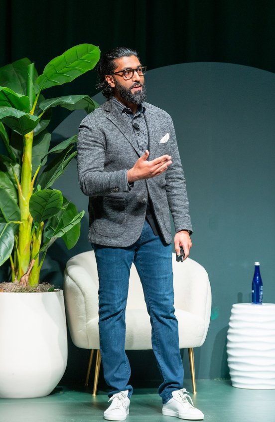 A bearded man in a gray blazer gesturing while speaking on stage, seated beside a potted plant and white armchair