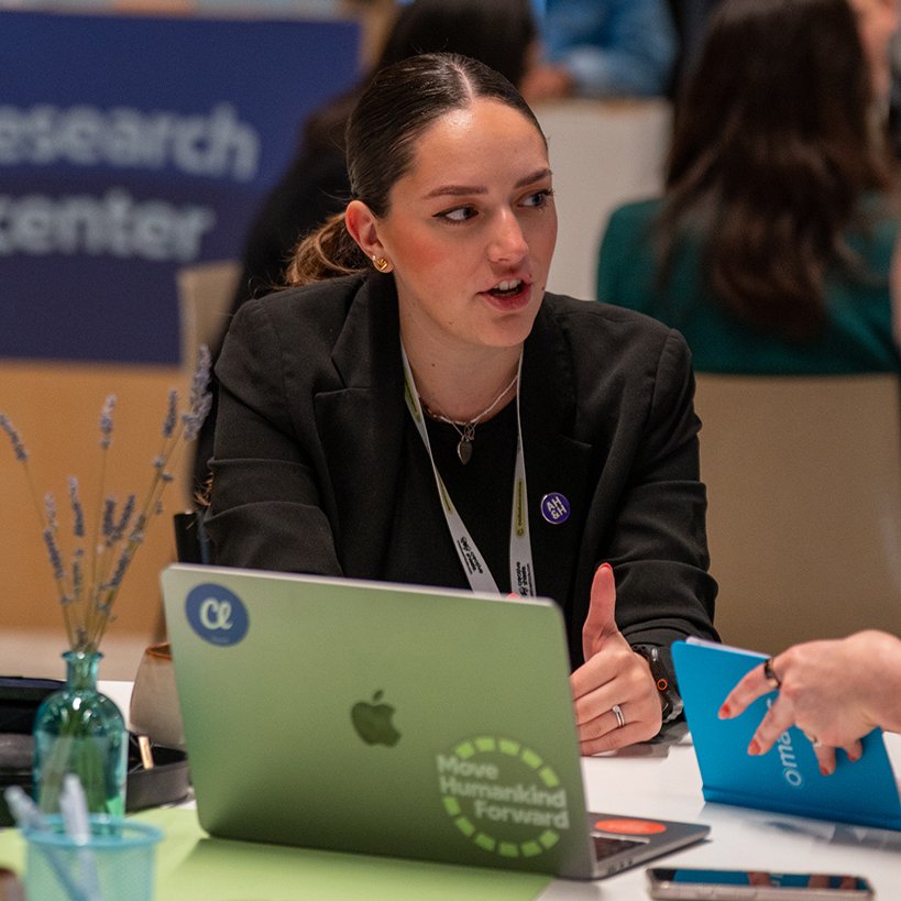 A woman in a black blazer talking with a colleague at a laptop during a conference networking session