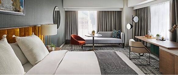 Hotel guest room with king bed, orange accent chair, round table, and workstation with skyline view